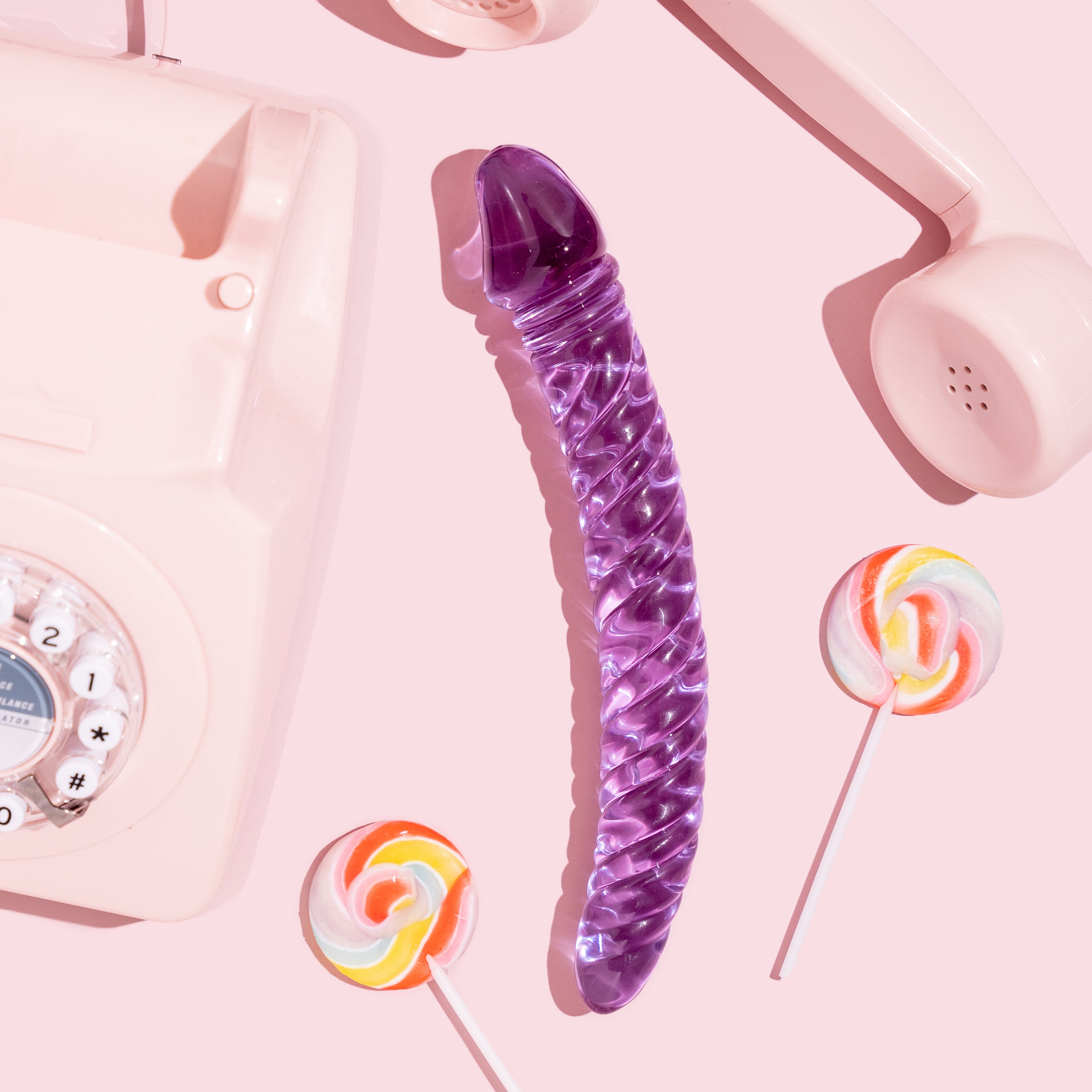 Curved purple glass ridged pleasure wand with thicker Fallic head and tapered end. Pictured on pink background with lollipops and a pink retro dial telephone.