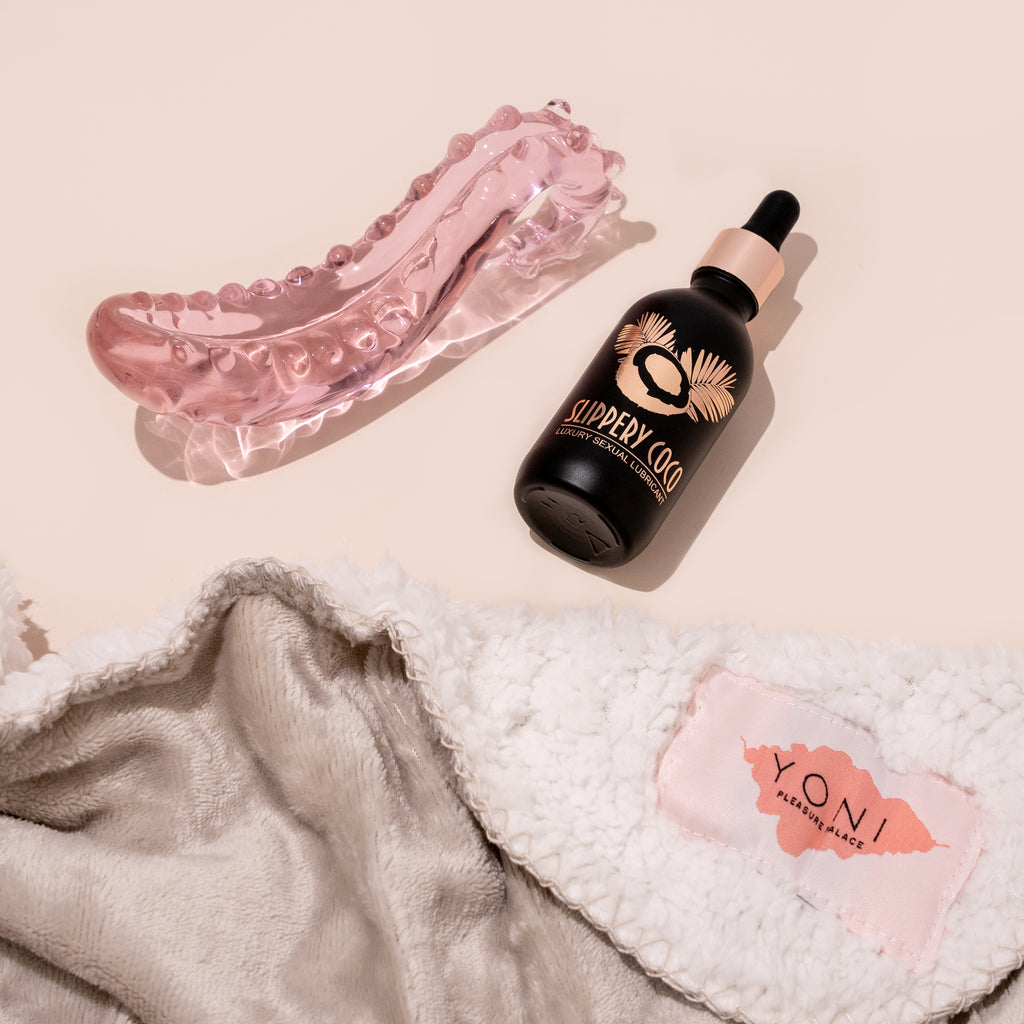 The squirt essentials kit containing a champagne coloured waterproof blanket, a bottle of slippery coco (coconut oil) lubricant in a black and gold bottle; and a pink glass sacred squirter pleasure wand. All are pictured on a beige background.