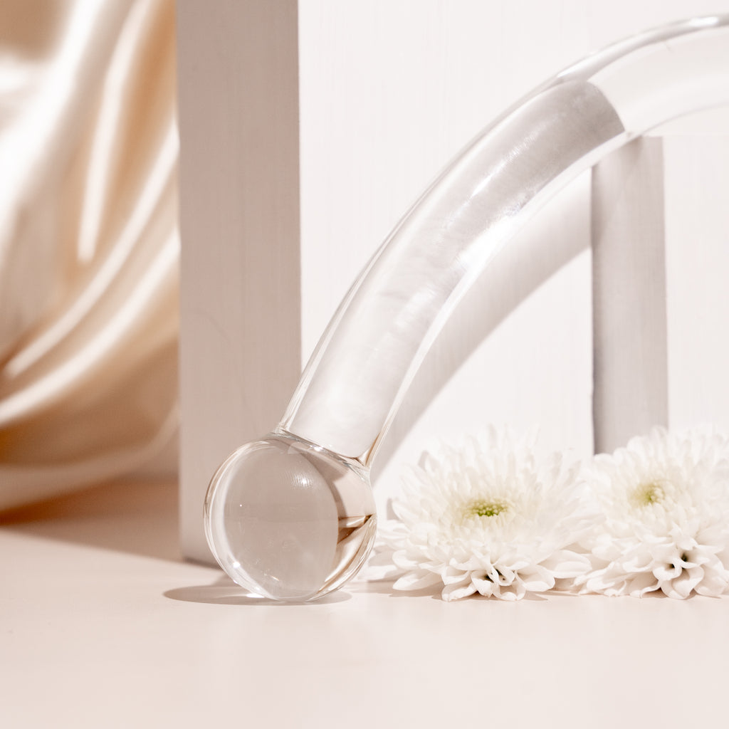 Curved clear glass pleasure wand with thicker spherical heads. Pictured on stone blocks with flowers and champagne coloured background.