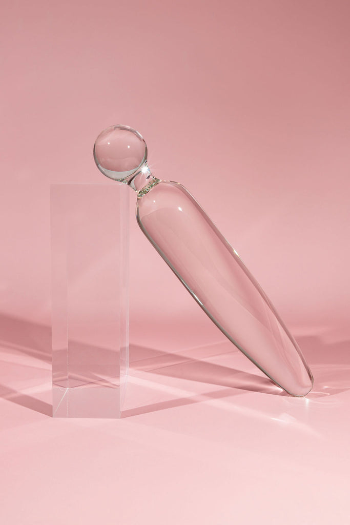 Clear glass pleasure wand with sphere shaped handle and traditional, straight end. Pictured on pink background with acrylic block.