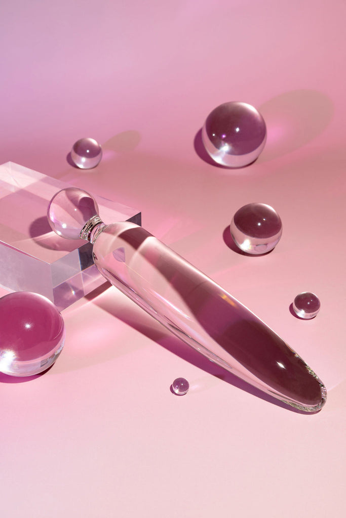 Clear glass pleasure wand with sphere shaped handle and traditional, straight end. Pictured on pink background with acrylic block and bubbles.