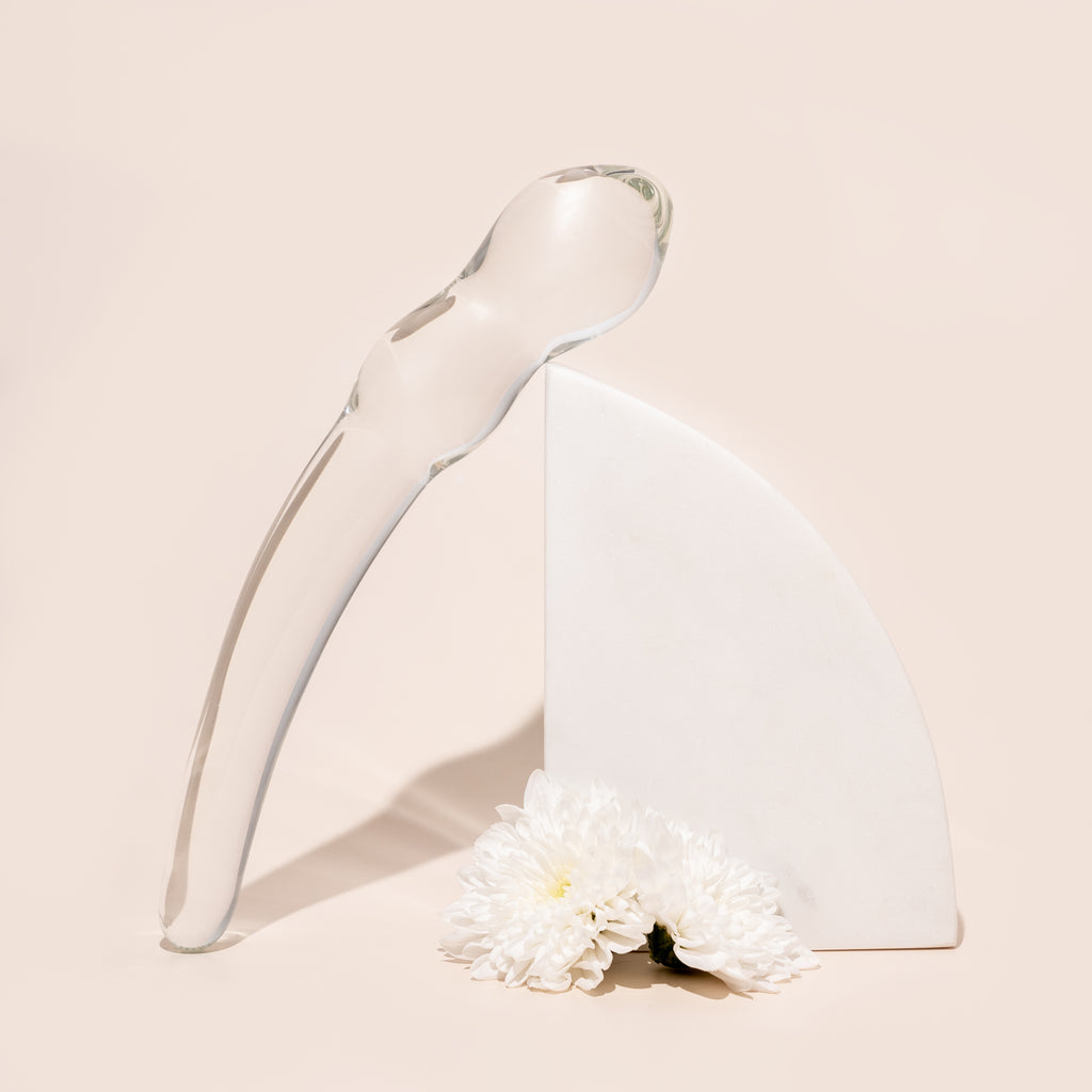 Curved clear glass pleasure wand with thicker head bobbles on one end and sleek tapered other end. Pictured on stone arch with flowers and white background.